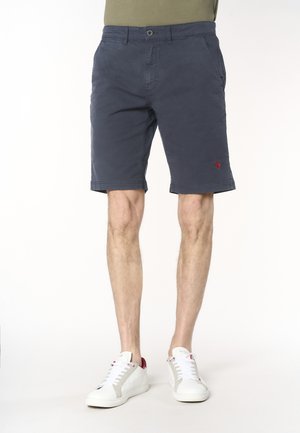 Uomo che indossa pantaloncini blu navy fino al ginocchio con tasche laterali e scarpe da ginnastica bianche con dettagli rossi, in piedi su uno sfondo uniforme.