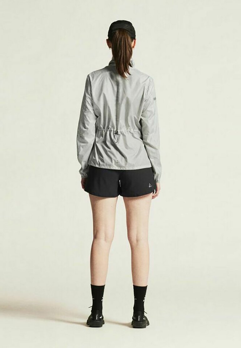 Veste coupe-vent gris clair avec une taille à cordon, portée avec un short noir. La veste possède des manches longues et un col montant.