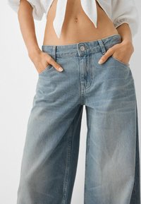 Kvinna i högmidjade vida blå jeans med händerna i fickorna och en knuten vit kort topp som visar en exponerad midja.