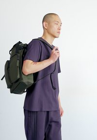 Olivgrön ryggsäck med flera fack, justerbara remmar, snygg design och matt yta. Bärs av en person i en lila outfit.