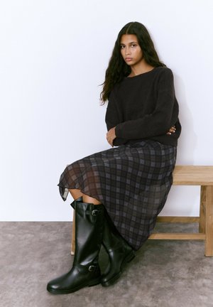 Botas negras de cuero hasta la rodilla con detalles de hebilla, llevadas con una falda de cuadros en tonos oscuros y un suéter texturizado en color oscuro.