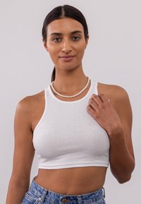 Witte geribbelde crop top met een aansluitend ontwerp en spaghettibandjes. Model draagt een parelketting en denimshorts, wat een casual stijl laat zien.