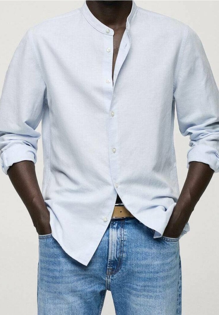 Camicia button-up in lino azzurro chiaro con colletto alla cinese, maniche lunghe arrotolate ai polsi, indossata con jeans blu e una cintura chiara.