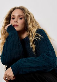 Maglione teal a trecce con polsini a coste, caratterizzato da una vestibilità rilassata e un motivo testurizzato, indossato da una persona che poggia il mento sulla mano.