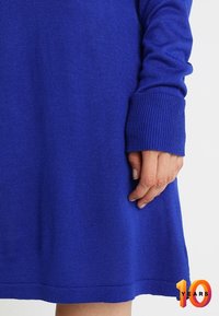 Person in einem langärmligen, königsblauen Strickkleid, wobei der Fokus auf der Hand und dem unteren Ärmel in der Nähe des Saums liegt.