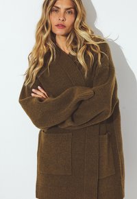 Blonde woman with wavy hair wearing oversized brown cardigan with pockets, standing with arms crossed against white background.