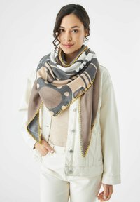 De sjaal heeft een geometrisch patroon met beige, grijze en witte kleuren, accentueert met gele stiksel langs de randen. De stof lijkt zacht te zijn.