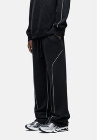 Zwart sweatshirt en joggingsbroek met witte accentstiksels, voorzien van een voorvak en een elastische tailleband met trekkoord, gecombineerd met grijze sneakers.