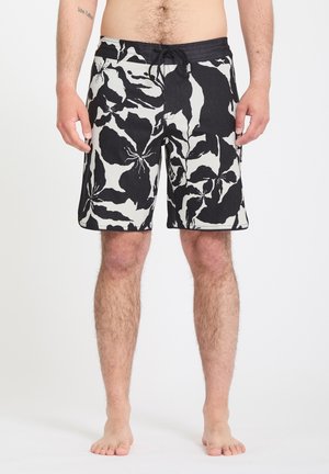 Adulte debout pieds nus portant un short de bain noir et blanc à motifs floraux avec ceinture à cordon.