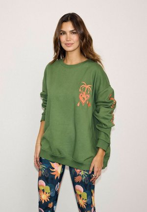 Woman wearing a green oversized sweatshirt with orange heart and palm tree design, paired with colorful patterned leggings.