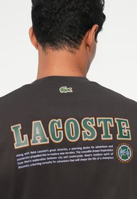 Juodos medvilnės marškinėliai su ryškiu "LACOSTE" tekstu žalia ir oranžine spalvomis. Mažas išsiuvinėtas krokodilo logotipas šalia kaklo. Minkšta tekstūra.