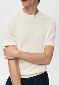 Camiseta de punto acanalado de color crema, de manga corta y diseño ajustado, con un cuello redondo. La tela parece suave y tiene una textura sutil.