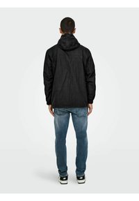 Jachetă neagră cu glugă din material ușor, cu o croială relaxată și manșete elastice, purtată cu blugi de denim bleumarin și adidași negri.