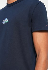 Μπλούζα από βαμβάκι σε ναυτικό μπλε χρώμα με λογότυπο που απεικονίζει βουνό σε πράσινο και λευκό, στρογγυλή λαιμόκοψη και κοντά μανίκια με μικρή διακριτική λεπτομέρεια της μάρκας.