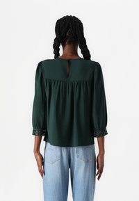 Blusa verde con una finitura texturizzata, maniche a sbuffo, schiena aperta con apertura a chiave e dettagli a taglio geometrico sulle maniche. Abbinata a jeans chiari.