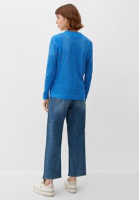 Frau mit kurzen braunen Haaren, die einen blauen, durchbrochenen Pullover, weite blaue Jeans und weiße Sneakers trägt und vor einem schlichten weißen Hintergrund steht.