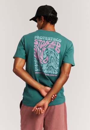 Petrolblauw katoenen T-shirt met een draaiende grafiek in roze en witte tekst op de achterkant. Model draagt rood geruite shorts en een zwarte pet.