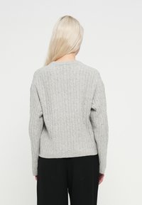 Világos hajú személy hátulnézetben, világosszürke kötött pulóvert és fekete nadrágot visel, sima fehér háttér előtt.