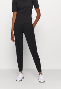 Schwarzes Sportoutfit mit tailliertem Kurzarmshirt und schmal zulaufenden Jogginghosen. Material wirkt glatt; Hosen haben gerippte Bündchen. Weiße Sneakers.