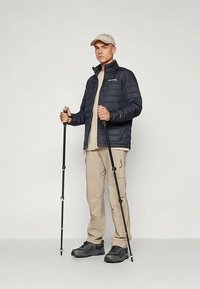 Jachetă puffer bleumarin, tricou bej și pantaloni, încălțări de drumeție negre și o pălărie ușoară. Ține bețe de trekking, stând pe un fundal neutru.