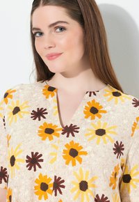 Blumenbluse mit einem hellbeigen Hintergrund, die gelbe und braune Blumenmuster aufweist, sowie einem V-Ausschnitt. Weicher Stoff.