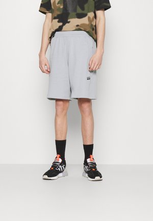 Pantaloni sportivi grigio chiaro realizzati in tessuto morbido, con vita elastica e tasche laterali, abbinati a una maglietta a stampa camouflage e sneakers nere.