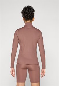 Haut à manches longues et col montant en mauve, fabriqué en tissu lisse et extensible. Associé à un short ajusté, montrant des poches sur les côtés.