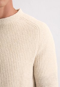 Beige Strickpullover mit gerippter Textur, ausgestattet mit einem runden Halsausschnitt und leicht versetzten Schulternähten, aus weichem, warmem Material.