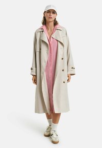 Cappotto lungo beige con grandi bottoni, indossato sopra un vestito rosa con bottoni, abbinato a sneakers bianche e un cappellino bianco. Texture lisce.