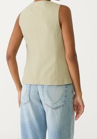 Ärmelloses, hellgrünes Oberteil mit glatter Textur und figurbetontem Design, kombiniert mit hellblauen Jeans, die über eine herzförmige Taschenstickerei verfügen.