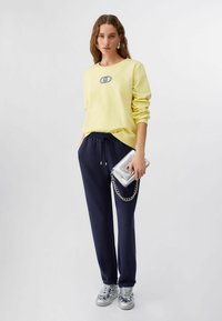 Femme portant un sweat-shirt jaune, un pantalon à cordon bleu marine, des baskets argentées, et tenant un sac à main argenté avec un détail en chaîne.