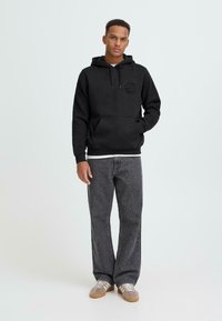 Juodas gobtuvas su kišene priekyje ir logotipu ant krūtinės, derinamas su pilkomis plačiomis džinsomis ir šviesiai rudais sportbačiais.