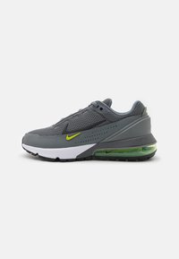 Zapatilla deportiva gris con parte superior de malla, plantilla blanca, suela de goma negra y amortiguación de aire verde distintiva. Acentos del logo en amarillo visibles.