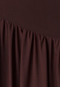 Tamno smeđa tkanina s finom mrežastom teksturom skupljena dijagonalno preko donjeg dijela, stvarajući mekane nabore i naboranost.