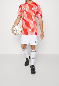 Persona vistiendo un traje de fútbol de Bayern Munich en rojo y blanco, sosteniendo un balón de fútbol Adidas de patrón estrella blanco, de pie con las piernas ligeramente separadas.
