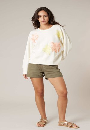 Femme portant un sweat-shirt blanc avec des imprimés de feuilles de palmier pastel, un short vert olive et des sandales beiges à brides, posant devant un fond uni.