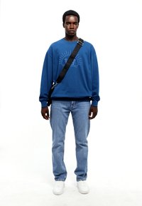 Blue sweatshirt with a sun graphic, paired with light blue jeans and white sneakers. Black strap bag worn crossbody.