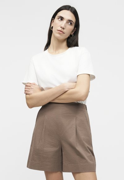 Frau mit langem dunklem Haar, die goldene Creolen, ein weißes Kurzarmshirt und hoch taillierte, strukturierte braune Shorts trägt, mit verschränkten Armen.