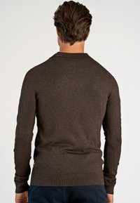 Man med mörkbrunt hår som bär en åtsittande mörkbrun långärmad tröja och marinblå byxor, vänd bort mot en enkel ljus bakgrund.