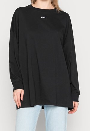 Femme portant un t-shirt noir à manches longues Nike surdimensionné et un jean bleu clair, debout devant un fond uni.