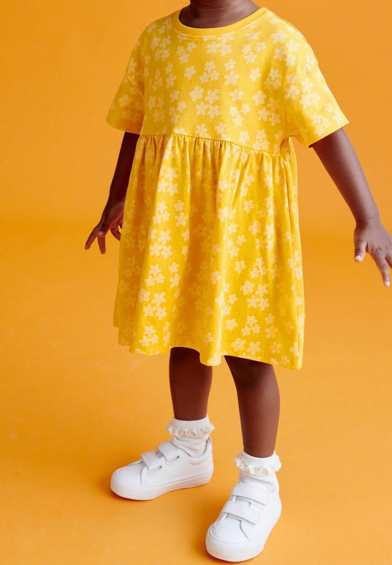 Enfant en robe fleurie jaune, chaussettes blanches à volants et baskets blanches à scratch, debout sur un fond orange.