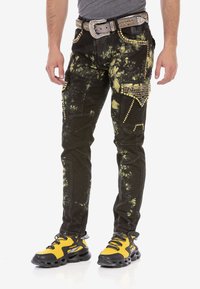 Schwarz-gelb gefärbte Jeans mit Batikmuster, verziert mit Nieten und Strasssteinen. Mit Taschen und einem dekorativen Gürtel ausgestattet. Ausführliche Nähte sind sichtbar.