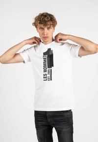 Camiseta blanca con texto impreso en negro "LES HOMMES" y un gran "1" con "EST. 2005." Mangas cortas y cuello redondo.