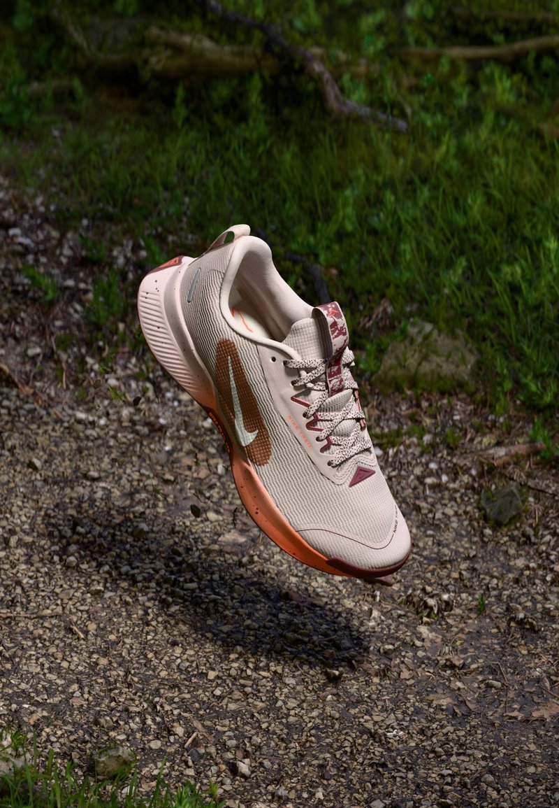Chaussure de course Nike rose clair et orange flottant au-dessus d’un chemin de gravier avec de l’herbe verte en arrière-plan.
