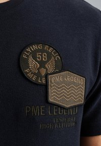 Mornarski košulja s teksturom koja sadrži dva vezena bedža: okrugli bedž s krilima i natpisom "FLYING RELIC 58" te heksagonalni bedž "PME LEGEND".