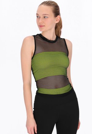 Femeie purtând un top negru fără mâneci din plasă peste un sutien sport neon galben, asortat cu colanți negri cu talie înaltă, uitându-se în lateral.