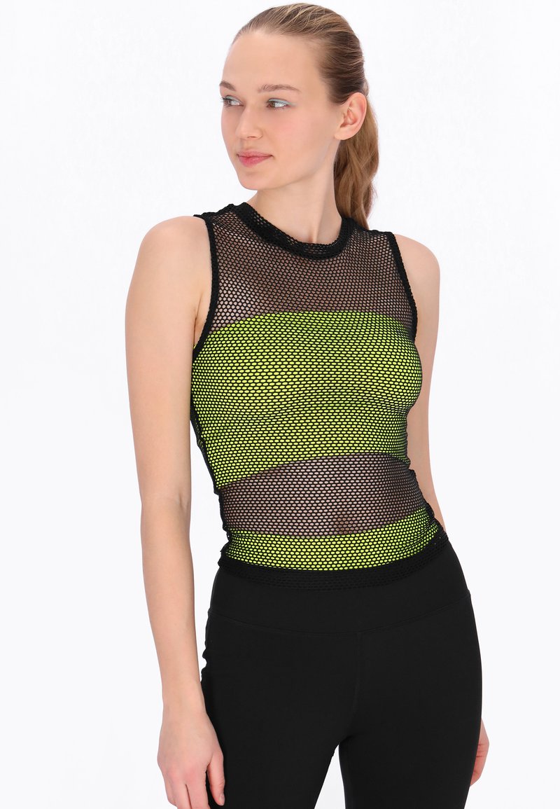 Kobieta ubrana w czarny bezrękawnik z siateczki na jaskrawoniebieskim sportowym staniku, w połączeniu z czarnymi legginsami z wysokim stanem, patrząca w bok.