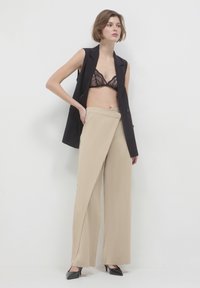 Donna che indossa pantaloni beige a gamba larga, un bralette in pizzo nero, un blazer senza maniche nero e scarpe con tacco a punta nere, in piedi contro una parete bianca.