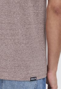 Camiseta marrón jaspeada hecha de tela suave, con corte recto, mangas cortas y una etiqueta de logo negra en el dobladillo.