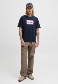 Fiatal férfi, hosszú hajjal és tetoválásokkal, sötétkék Jack & Jones pólót, barna nadrágot, bézs cipőt és szürke sapkát visel, egy fehér háttér előtt áll.